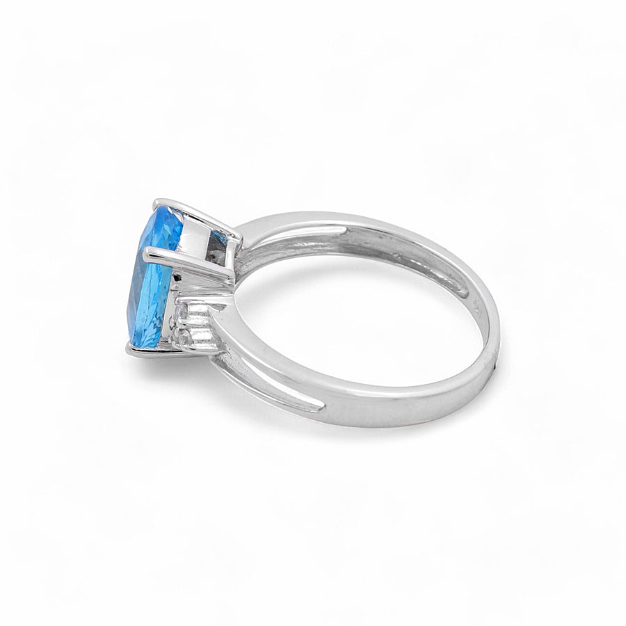 The 14K White Gold Women's Fashion Ring by Miral Jewelry, featuring an oval blue topaz and two small diamonds on the band, is viewed from the side against a white background. Its design is reminiscent of a contemporary diamond ring.