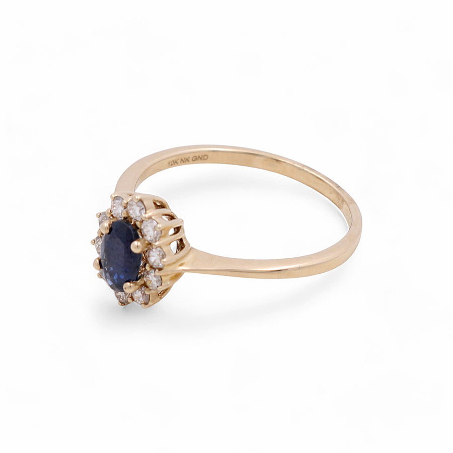 The Miral Jewelry 14K Yellow Gold Woman Ring with Diamonds and Sapphire features an oval blue gemstone surrounded by small clear stones in a floral pattern, making it a stunning piece of jewelry.