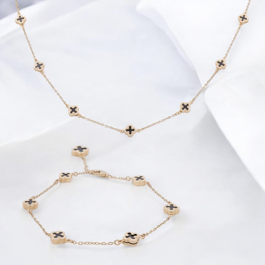 A 14K yellow gold necklace and bracelet set from Miral Jewelry, featuring clover-shaped accents with onyx, elegantly displayed against a white fabric background.