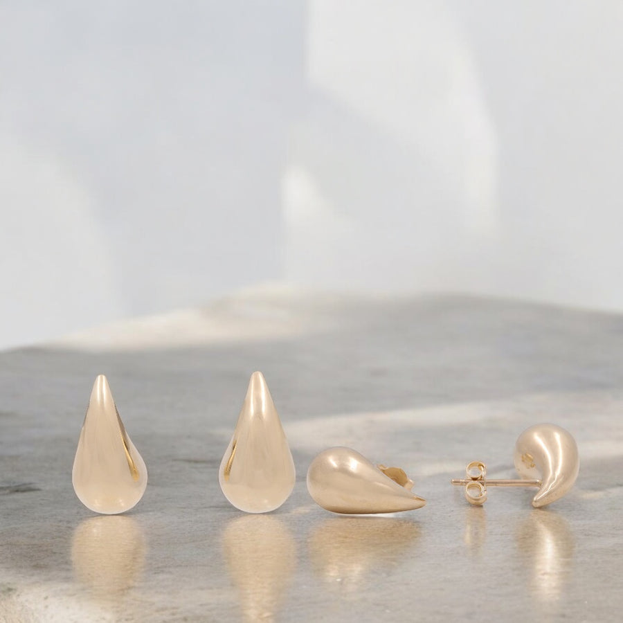 Displayed on a smooth surface is a set of Miral Jewelry's 14K Yellow Gold Small Drop Earrings. Two earrings stand upright, while the other two are laid flat, elegantly showcasing both their front and side views.