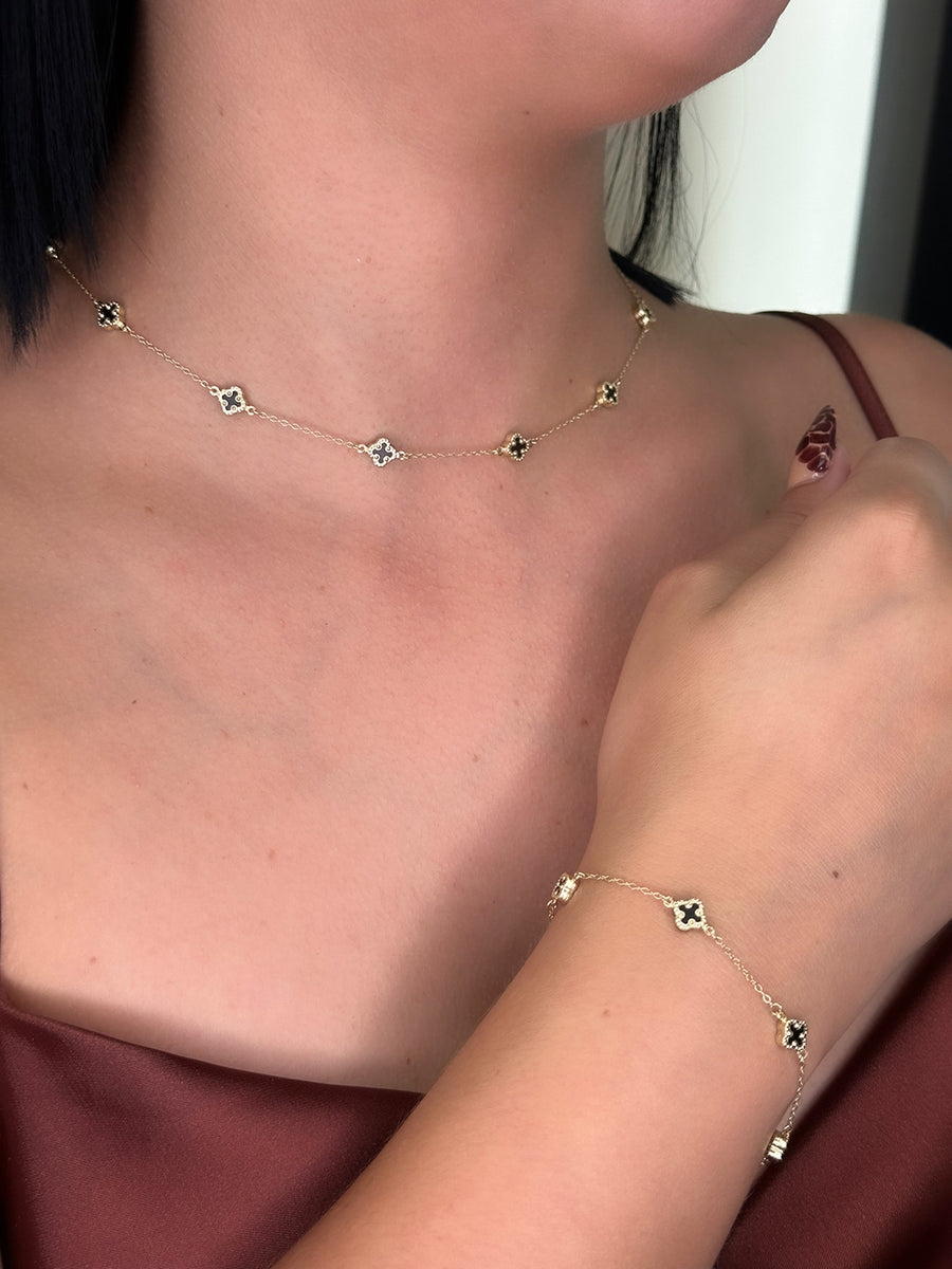 Person wearing the 14K Yellow Gold Fashion Clover with Onyx Set by Miral Jewelry, featuring a gold necklace and matching bracelet with small black clover-shaped onyx pendants, shown in close-up against a maroon outfit.