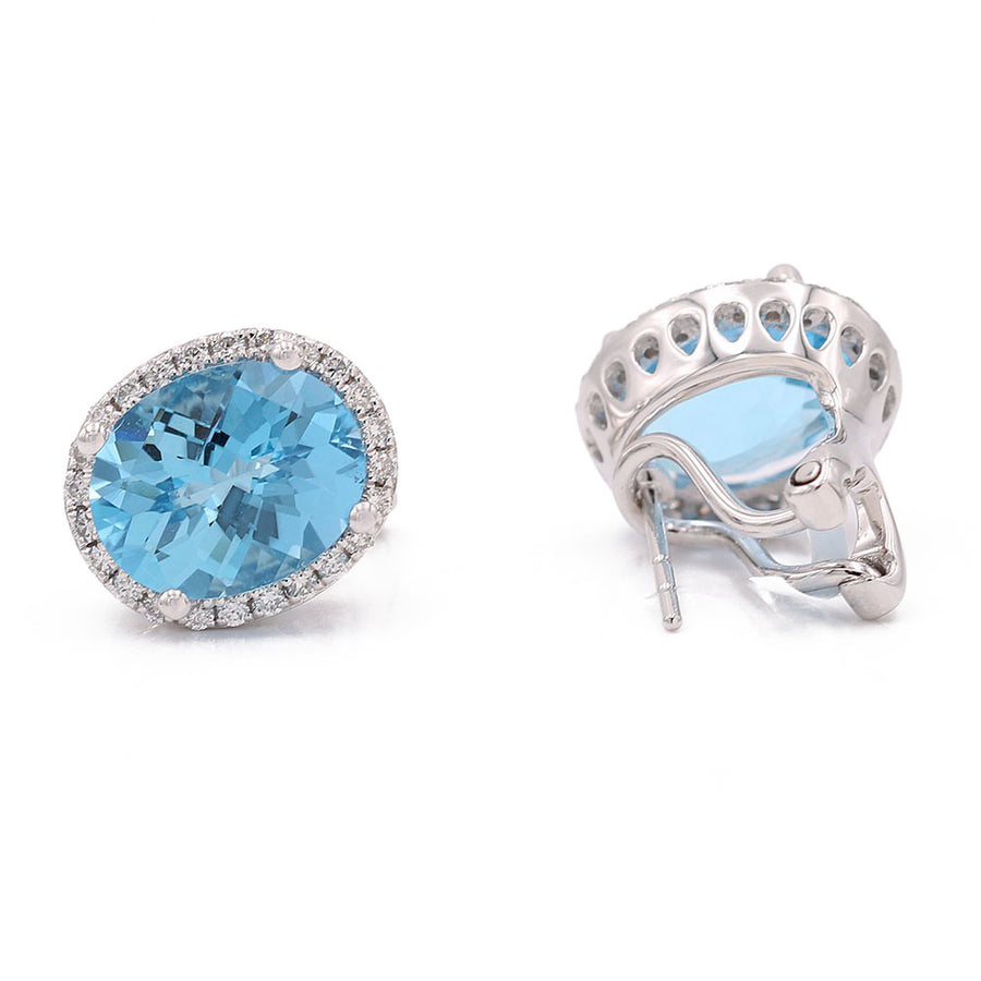 Pair of Miral Jewelry White 14K Karat Stud Omega Clasp Earrings featuring large oval blue topaz gemstones surrounded by small round diamonds. One earring is shown from the front, and the other is displayed from the side.